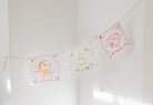 Three decorative square pennants strung along a thin cord, hanging against a white wall. The center pennant reads “5 Cinco” with green and orange lettering, while the side pennants feature pink and orange illustrations with Spanish text.