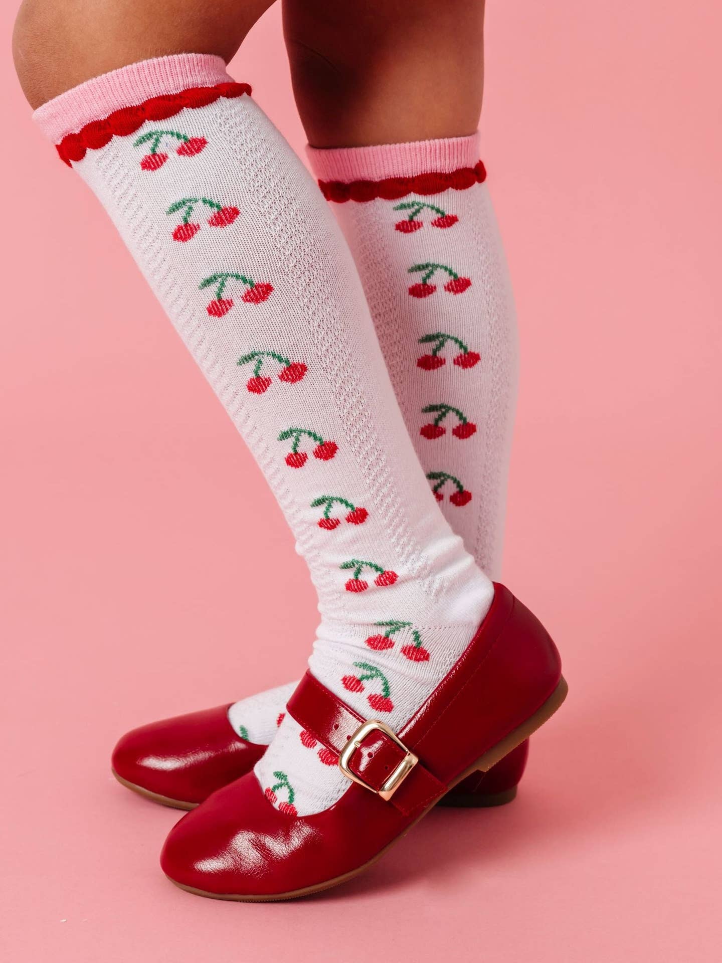 White knee-high socks with cherry pattern worn with red shoes on a pink background