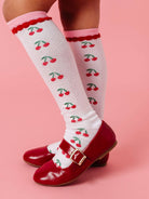 White knee-high socks with cherry pattern worn with red shoes on a pink background