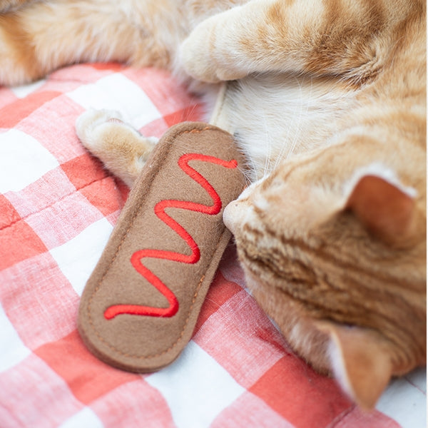An orange tabby cat lies on a red-and-white check blanket next to a brown felt toy shaped like a hot dog with red embroidered mustard lines.