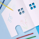 A white house-shaped children's drawing board with window and arch cutouts, shown on a light blue background with colored pencils around it. The board has simple hand-drawn flower and squiggle marks in blue, pink, green, and gray.