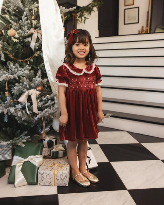 A young girl stands in front of a decorated Christmas tree wearing a festive red velvet dress with white lace trim and gold bead details. She is posed on a black and white checkered floor next to wrapped gifts, with a staircase in the background.