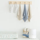 A light wood wall-mounted hook rack with four hanging fabric accessories in soft neutral and blue tones, displayed in a bright nursery-like setting above a changing surface with baby textiles and a white humidifier.