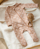 A baby footed sleeper in a dusty pink floral print with a front zipper, laid flat on a cream textured blanket next to a small woven rattleskin rattle.