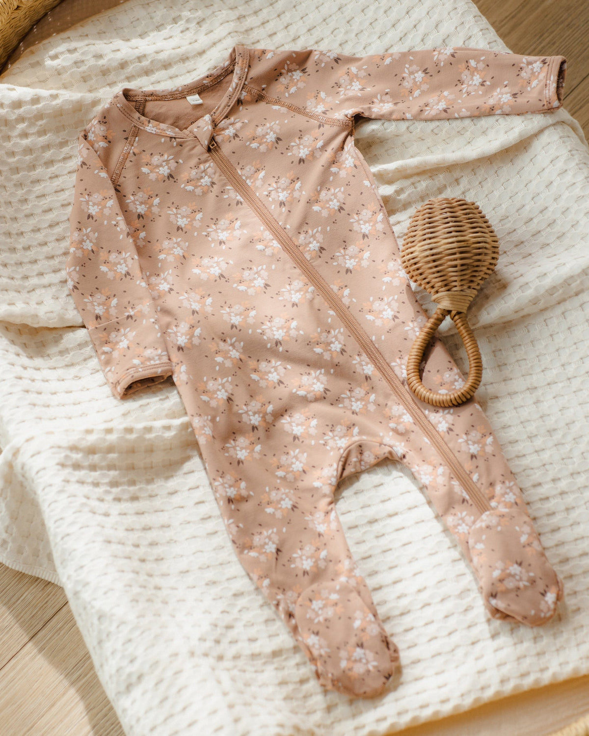 A baby footed sleeper in a dusty pink floral print with a front zipper, laid flat on a cream textured blanket next to a small woven rattleskin rattle.