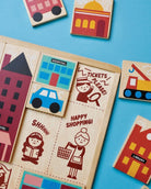 A close-up, top-down view of the Mentari Wooden Town Puzzle showing colorful, chunky wooden puzzle pieces partially placed in their tray, featuring buildings, a blue car, and illustrated characters with text reading "Tickets Please," "Shhhh!," and "Happy Shopping!"