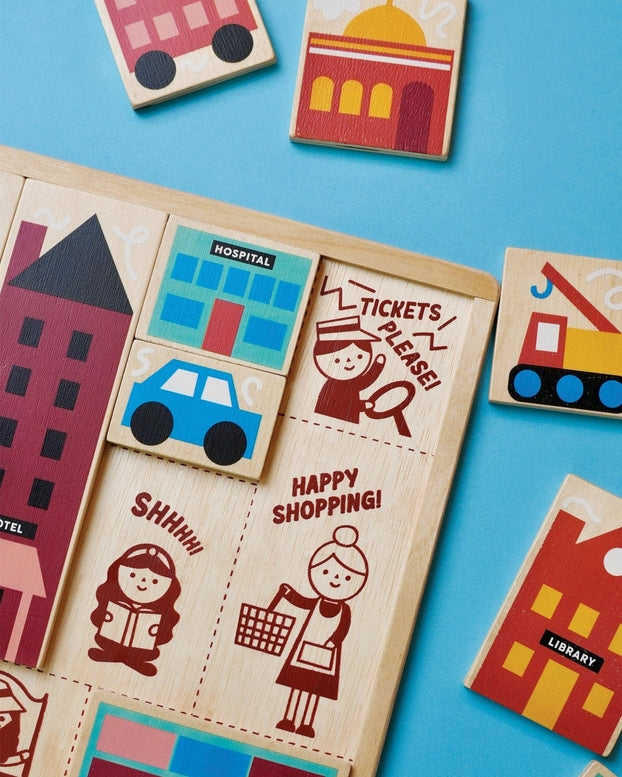 A close-up, top-down view of the Mentari Wooden Town Puzzle showing colorful, chunky wooden puzzle pieces partially placed in their tray, featuring buildings, a blue car, and illustrated characters with text reading "Tickets Please," "Shhhh!," and "Happy Shopping!"