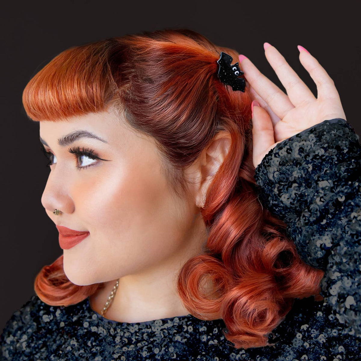 A close-up portrait of a woman with retro-style red hair, wearing a black sequined top and a small black hair clip, set against a dark background.