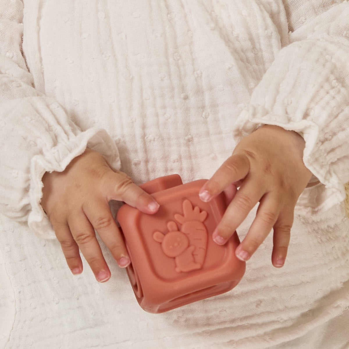 Child's hands holding a pink silicone sand mold with a paw print design against a white textured background.