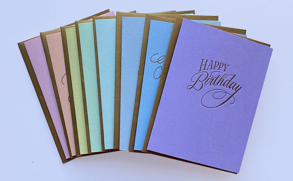 A fan of colorful greeting cards with gold envelopes, featuring a prominent lavender purple card with 'Happy Birthday' written in gold script.