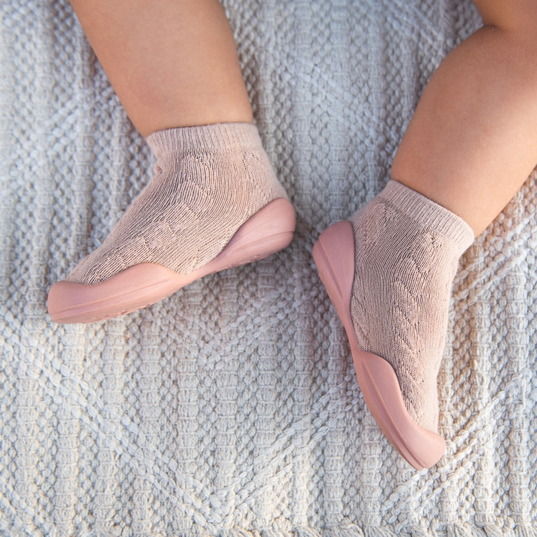 A close-up of a baby's feet wearing pale pink knit socks with flexible, wedge-shaped soles, resting on a textured cream knit blanket.