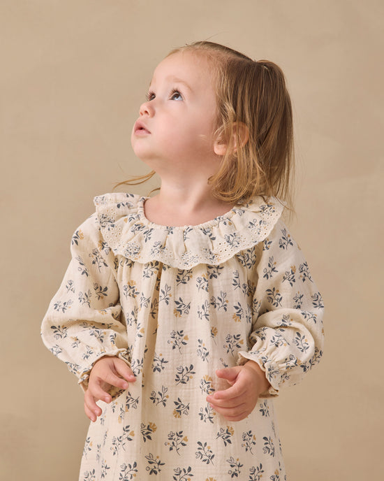 A young girl with light brown hair in a ponytail wearing a cream-colored long-sleeve dress with a delicate blue and yellow floral print, looking upwards against a warm beige background.