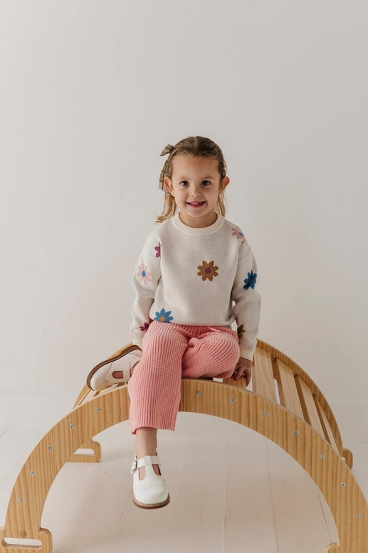 A young girl with braided hair sits on a wooden arched bench, wearing a cream knit sweater with colorful embroidered flowers, pink ribbed trousers, and white shoes against a plain light background.