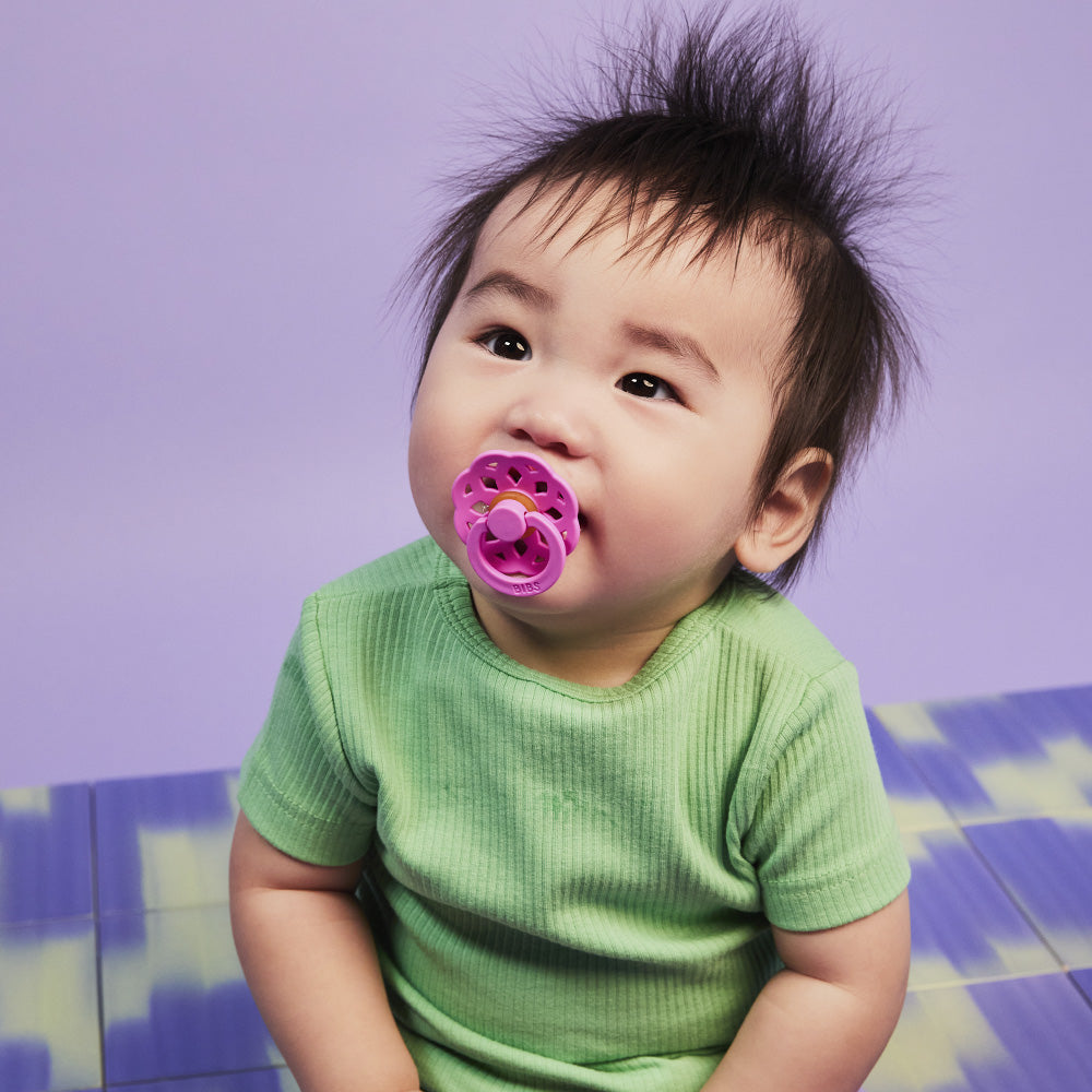 Baby with a purple pacifier wearing a green shirt against a purple background