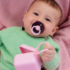 Baby in a green sweater with a purple pacifier holding a pink cup against a pink background