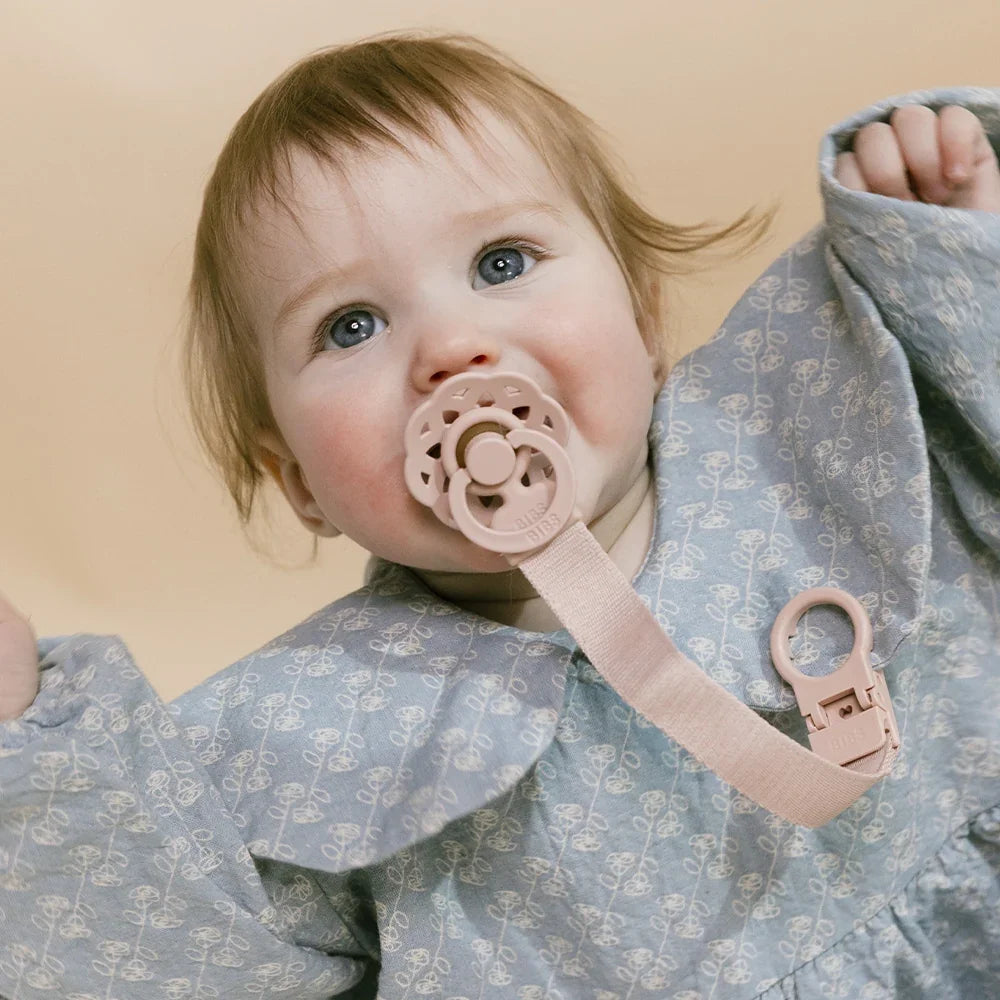 A close-up of a baby wearing a dusty blue patterned dress and holding a pale pink pacifier clip with a pacifier in their mouth, set against a solid beige background.