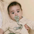 Baby wearing a beige outfit with a pacifier held by an adult