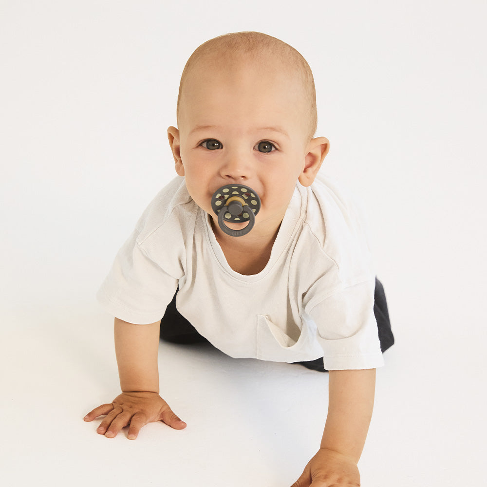 Baby wearing a white onesie with a black and gold pacifier on a white background