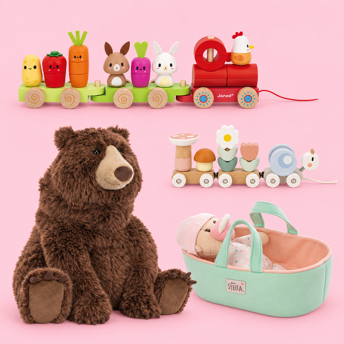 A collection of children's toys arranged on a pink studio background: a large brown plush bear, a red toy train with colorful vegetable and animal figures, a small wooden pull toy train, and a mint green baby bassinet with a doll inside. The bassinet label reads Stella.