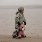 A child wearing a light sage green hooded rain suit, black gloves, and a gray cap stands on a sandy shore holding a pink teddy bear against a misty body of water.