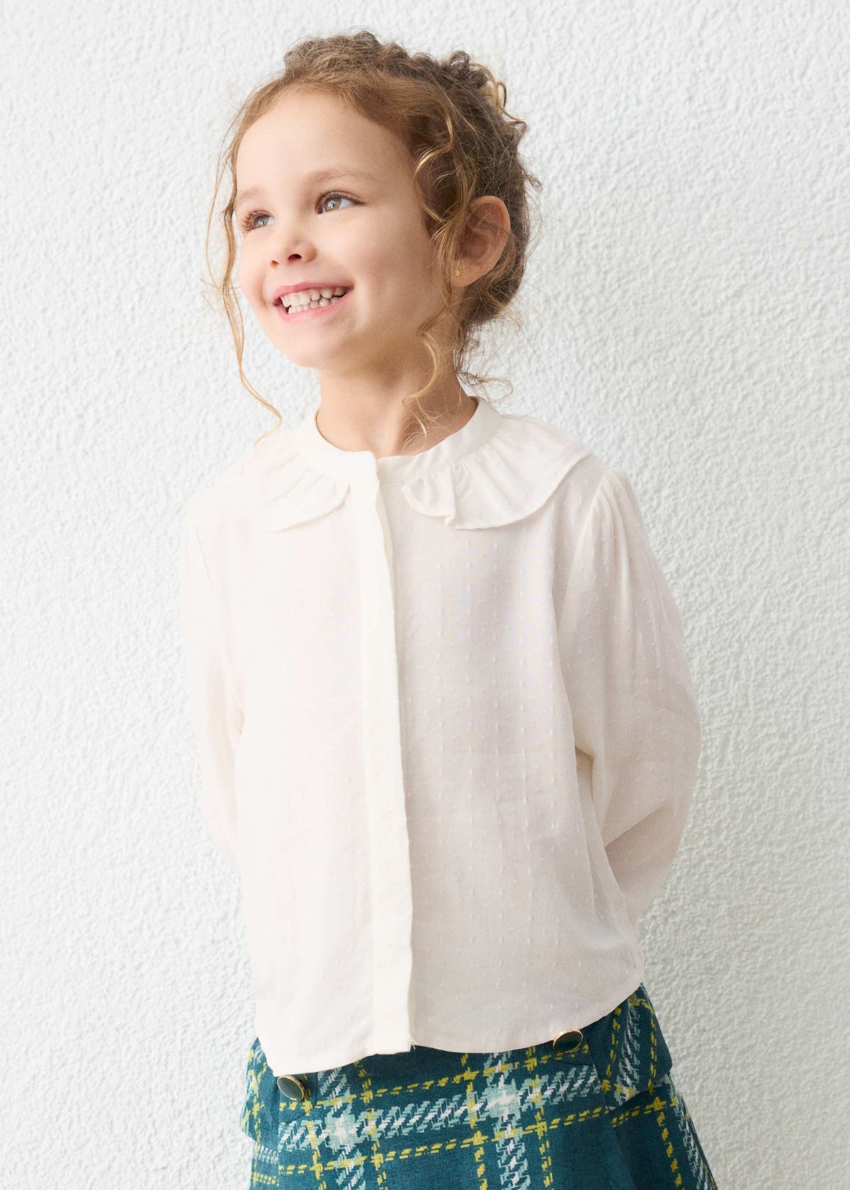 Young girl wearing a white blouse and plaid skirt against a white wall
