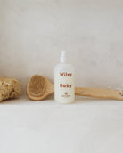 A bottle of Wiley Baby baby shampoo stands on a light surface against a textured off-white wall, styled with a wooden brush and a natural sponge.