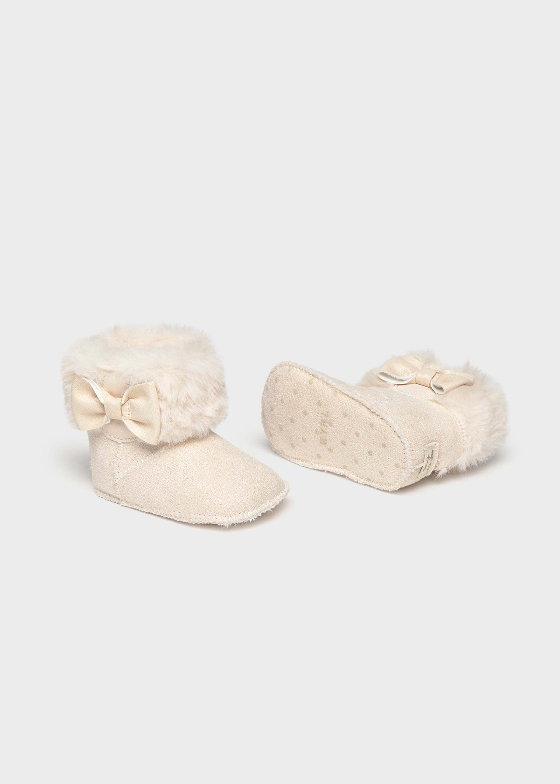 A pair of cream-colored baby booties with fluffy faux-fur trim and bow details, shown on a light gray background.