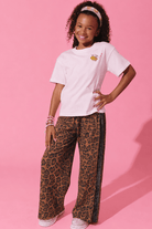 A young girl modeling a white t-shirt with a small yellow graphic and brown leopard-print wide-leg pants with black side stripes, standing against a solid pink background.