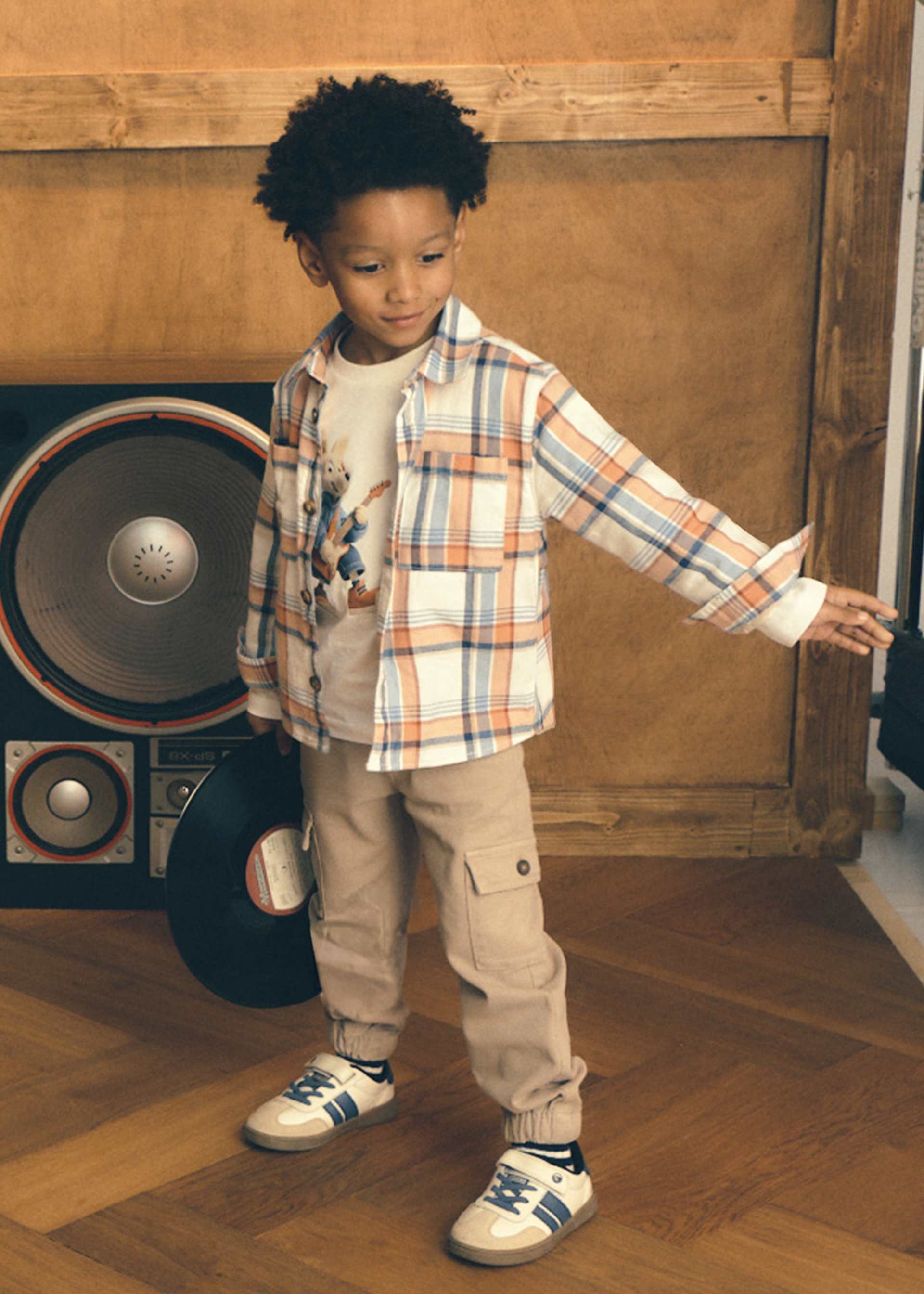 A young boy wearing a plaid button-up shirt, beige cargo pants, and white sneakers stands in a room with wood paneling and a large speaker behind him.