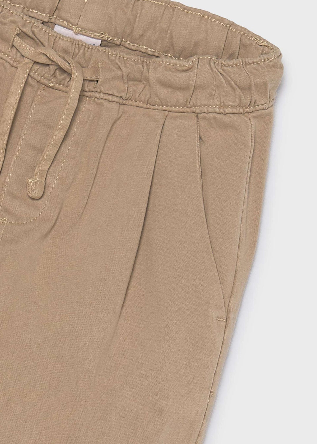 Close-up of a pair of pleated trousers in a light tan or khaki color, showing the elastic waistband, drawstring, and side pocket stitching against a light gray background.