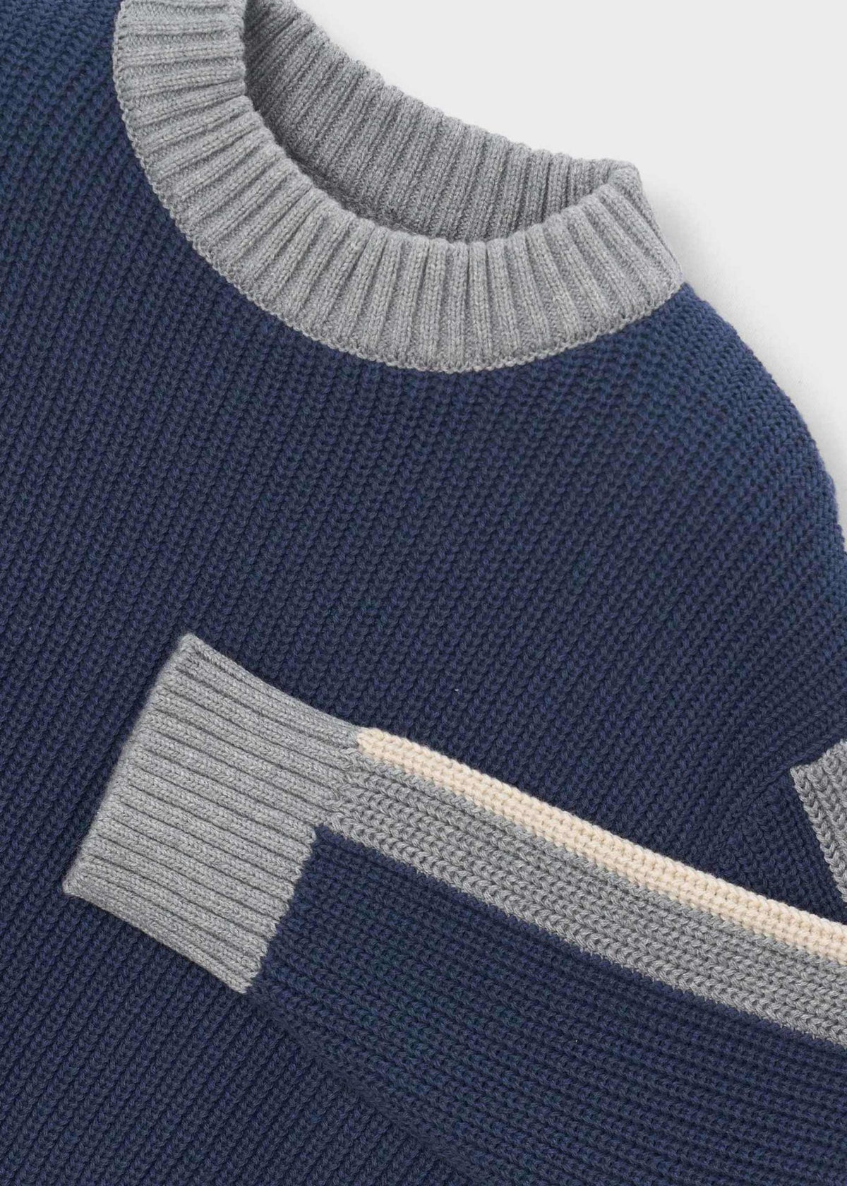 Close-up of a ribbed knit sweater in navy blue with light gray ribbed trim at the crew neck and sleeve cuff, plus a contrasting light beige stripe running diagonally.