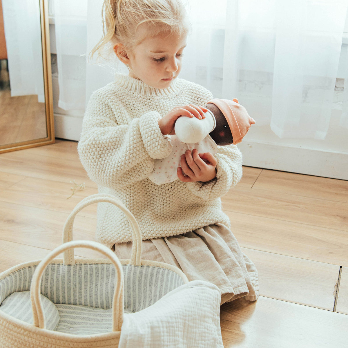 Child in a white sweater holding a toy bottle on a wooden floor.