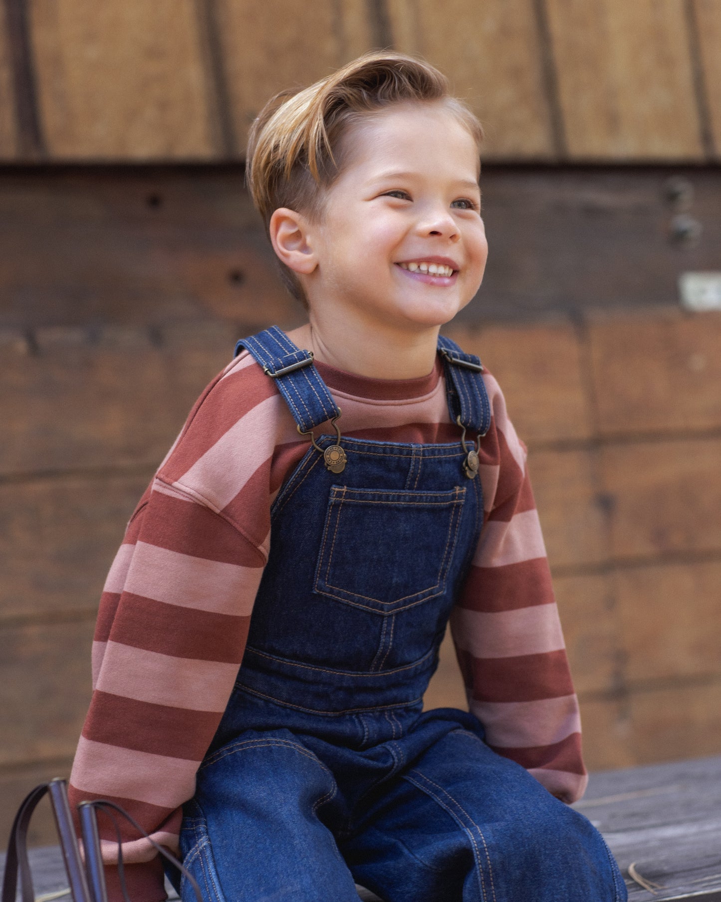 A young boy smiling and looking to the side, wearing a brown and pink striped long-sleeve shirt under blue denim overalls, sitting outdoors against a rustic wooden wall.