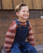 A young boy smiling and looking to the side, wearing a brown and pink striped long-sleeve shirt under blue denim overalls, sitting outdoors against a rustic wooden wall.