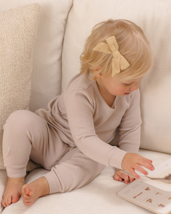 A toddler with blonde hair and a beige bow sits on a cream sofa, wearing a matching ribbed long-sleeve top and pants set in a light beige/taupe color. The child is interacting with a book.