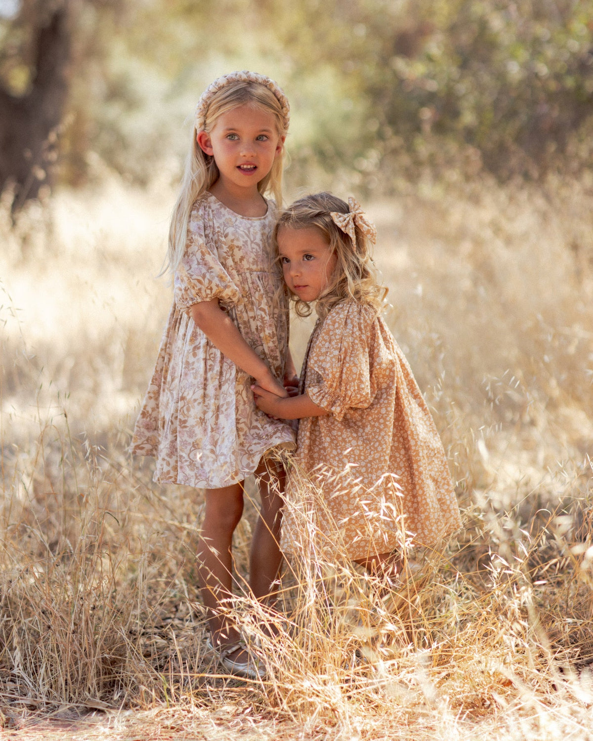 Two young girls standing in a field of dry golden grass, wearing matching beige and white floral print dresses with short puff sleeves and headbands.