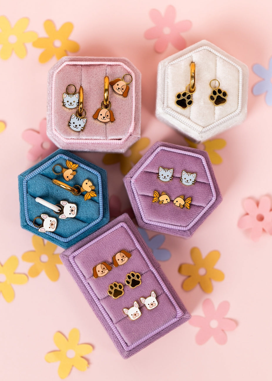 A styled flat-lay of five hexagonal jewelry boxes in pink, white, blue, and purple, each holding small gold-tone animal-themed earrings. The earrings feature cat and dog faces, paw prints, and bows, arranged on a pastel pink background with scattered paper flowers.