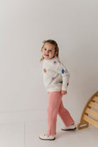 Child wearing a floral sweater and pink pants standing against a plain background