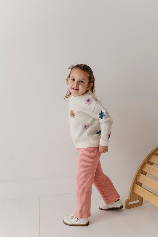 Child wearing a floral sweater and pink pants standing against a plain background