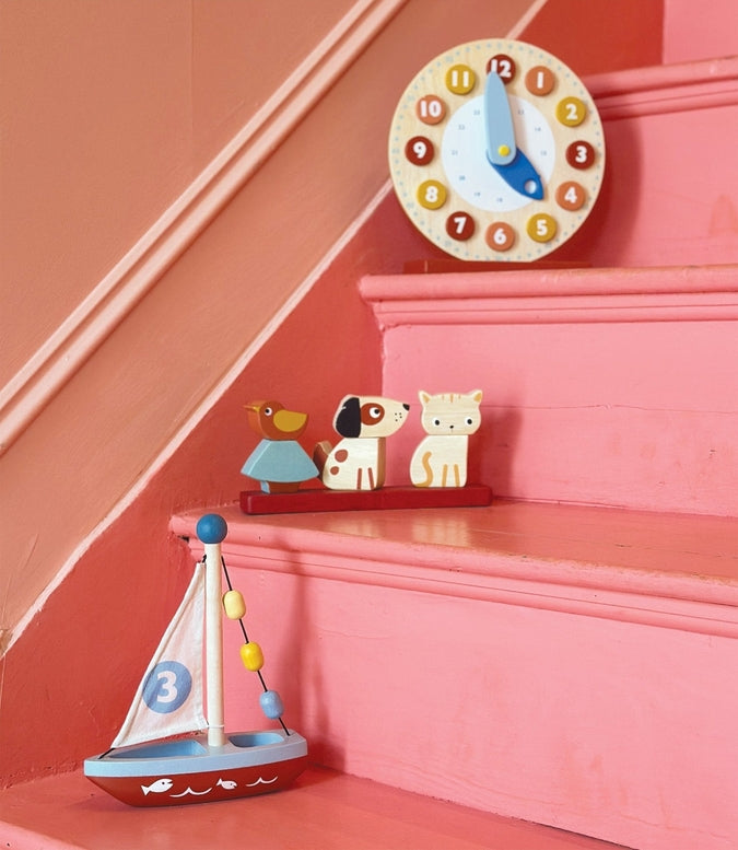 Wooden Pet Stacker toy by Mentari featuring a rectangular red base holding three removable pieces: a spotted white and brown puppy on the left, a blue and yellow bird on a tree in the center, and a white kitten with orange markings on the right, resting on a pink staircase beneath a wall-mounted blue and white clock.