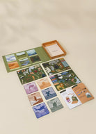 A children's card game set arranged on a warm beige background, including illustrated cards, a green play mat, an orange storage box, and instruction sheets.