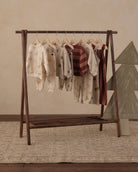 A wooden A-frame clothing rack holding several children's long-sleeve tops and shorts in cream, beige, and burgundy tones, styled in a warm neutral room with a patterned rug and a decorative tree on the right.