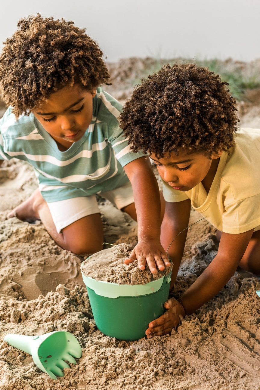 Two children playing with sand and a green bucket on a sandy beach.