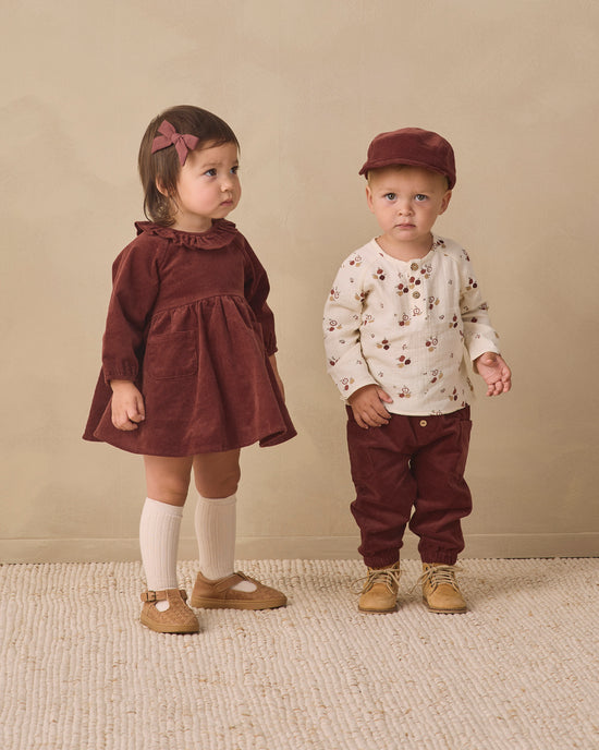 Two toddlers standing in a studio setting. The girl wears a dark burgundy corduroy dress with white knee-high socks and tan shoes, and the boy wears a cream long-sleeve shirt with a small floral print, burgundy pants, tan shoes, and a burgundy cap.