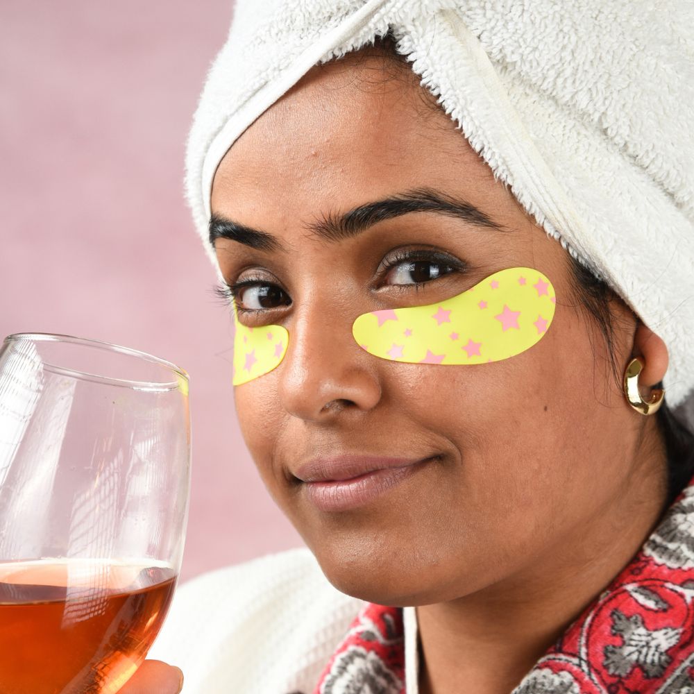 Woman with eye patches holding a glass of wine, wearing a towel on her head.