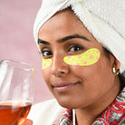 Woman with eye patches holding a glass of wine, wearing a towel on her head.