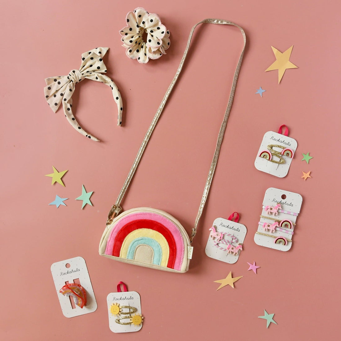 Children's accessories including a rainbow purse, headband, and hair clips on a pink background.