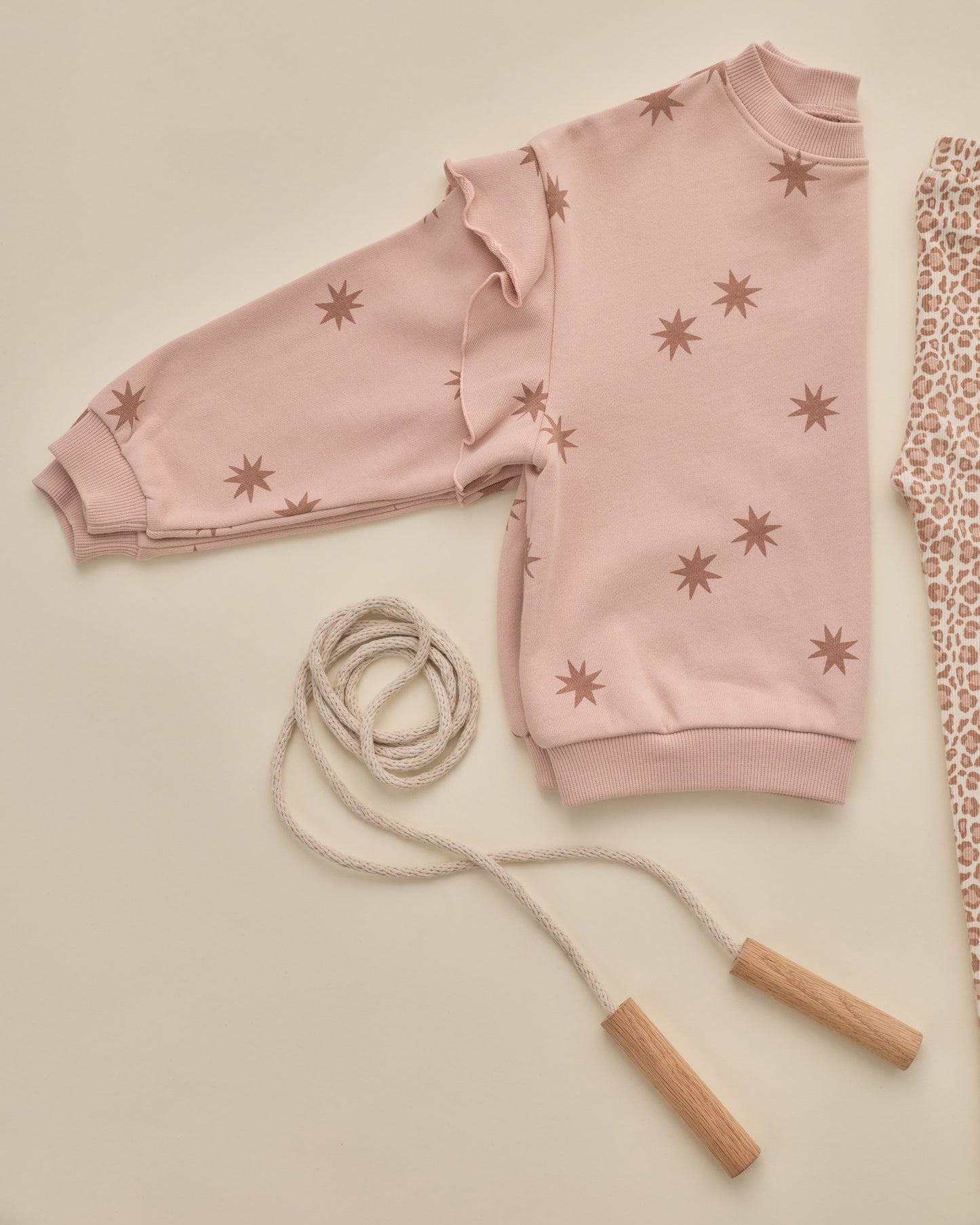 A light pink long-sleeve sweatshirt with brown starburst prints, styled in a flat lay with a beige rope jump rope and a patterned fabric on a warm cream background.