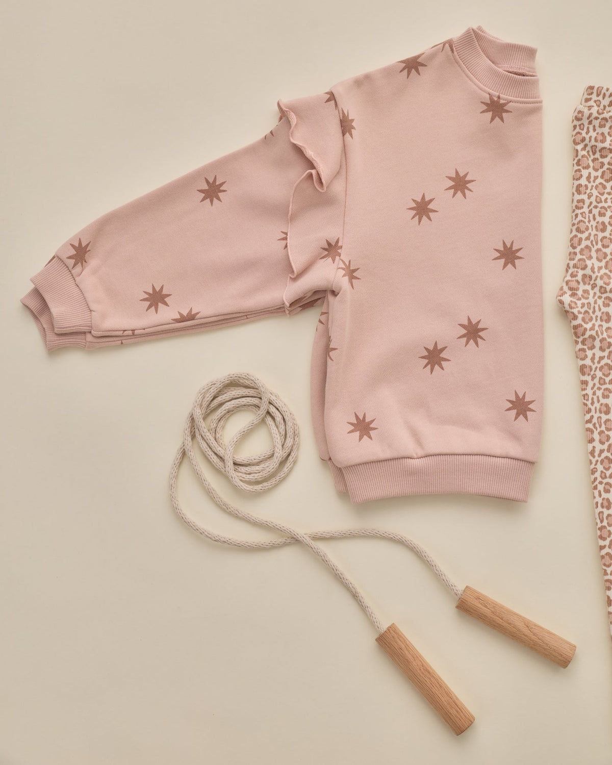 A light pink long-sleeve sweatshirt with brown starburst prints, styled in a flat lay with a beige rope jump rope and a patterned fabric on a warm cream background.