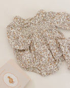 A baby long-sleeve romper with a smocked chest and ruffled collar, featuring a delicate beige and brown floral print on a cream background, styled flat-lay on a light cream surface next to a book.